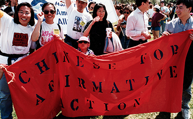 Image description: CAA friends, allies, and staffers attend a 1996 protest in opposition to Proposition 209, which banned affirmative action in the state of California. They are holding up a red banner that reads "Chinese for Affirmative Action".  