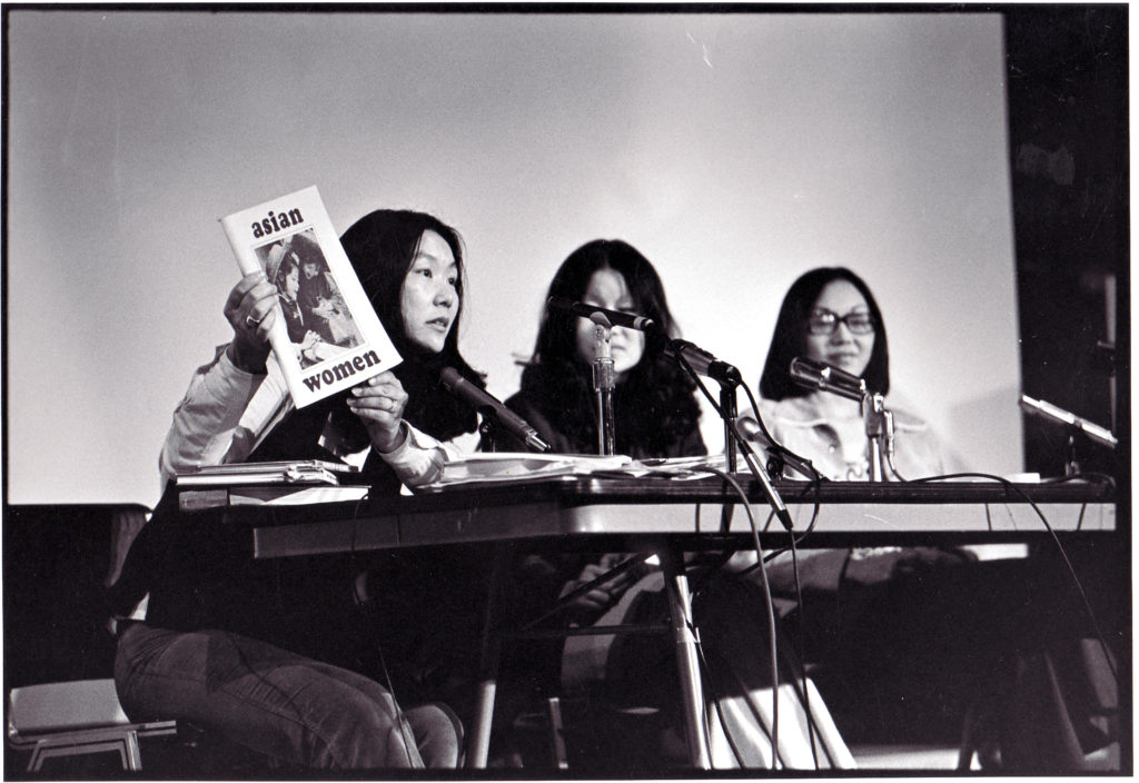 Image description: Chinese American women representing CAA at a 1970's community event. They are sitting at a table with microphones affixed to one end. The woman on the left is holding a copy of a magazine with the title "Asian Women."   