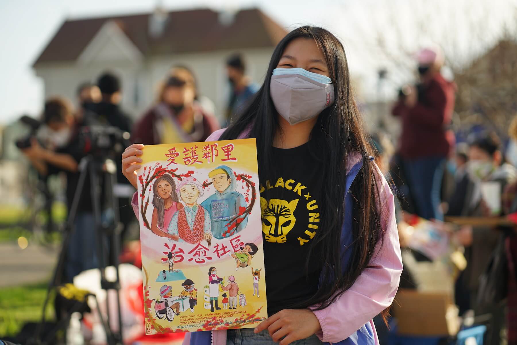 A young Asian woman with long hair holds up a poster depicting three people and a heart around them. She is wearing an Asian-Black Unity T-shirt.