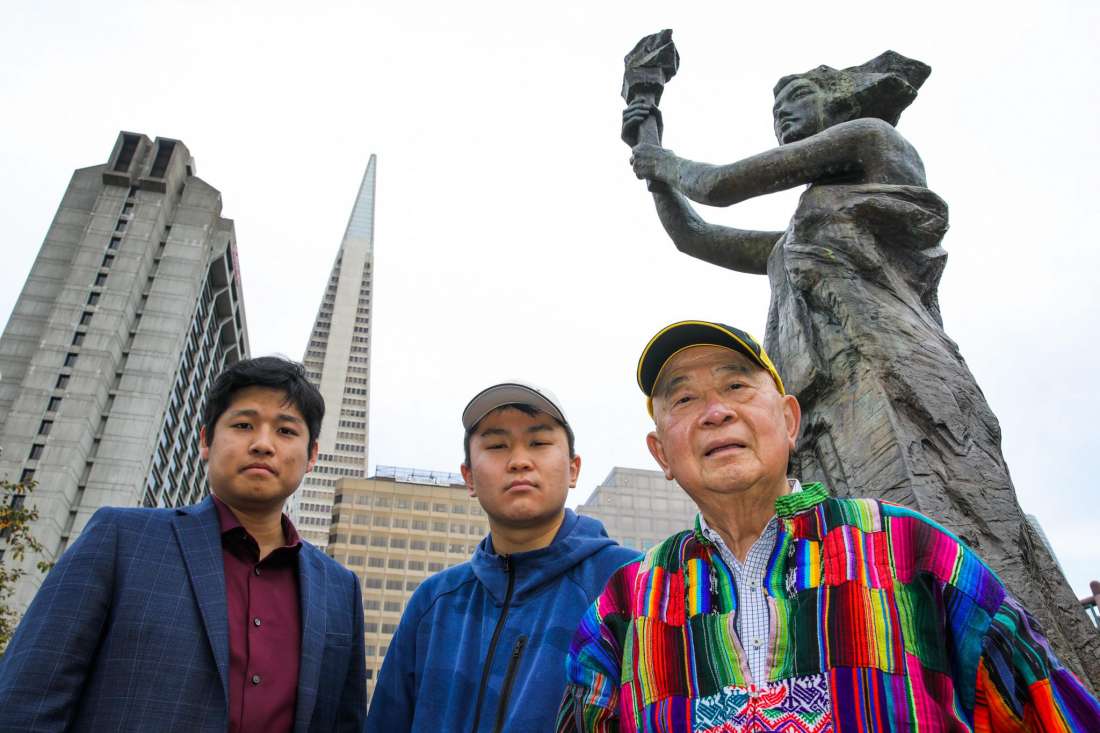 Three Asian people, two young, one older, all male-presenting stand in front of a statue.