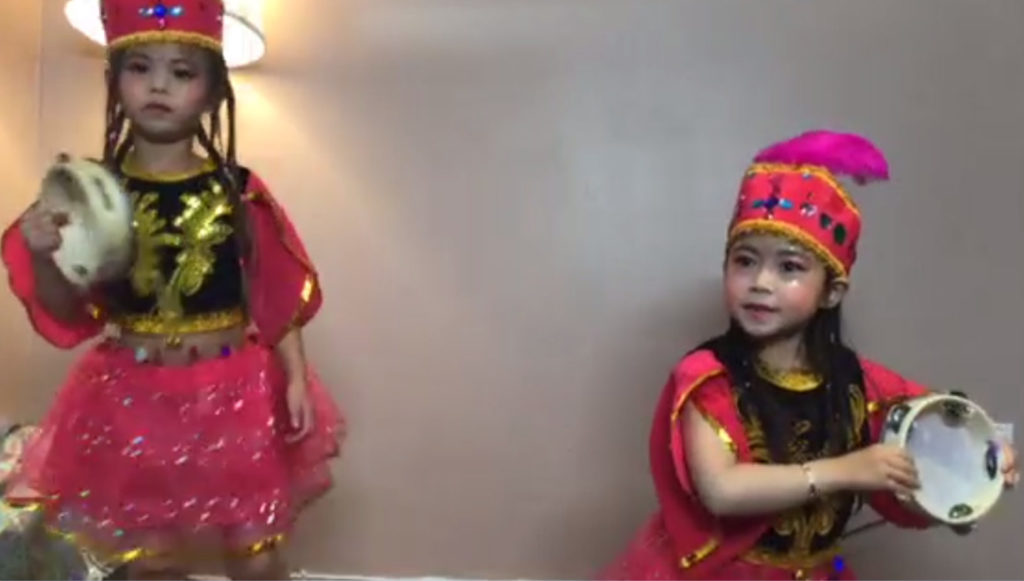 Two East Asian girls, wearing red costumes, perform a dance. They are holding tambourines.