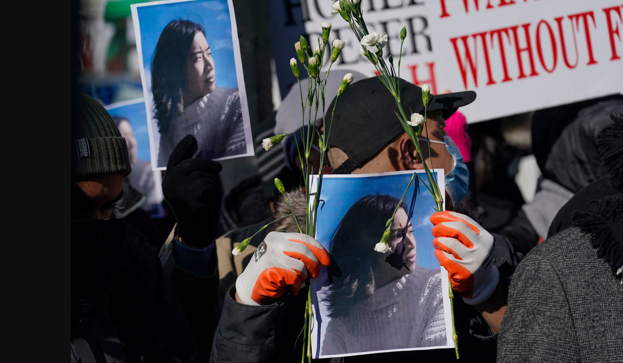 A protestor in a black hat and gloves holds up a photo of Christina Yuna Lee who was murdered in her apartment in New York City. Flowers are affixed to the photo. Other photos and protest signs can be seen in the background. Photo credit: Seth Wenig, Associated Press.