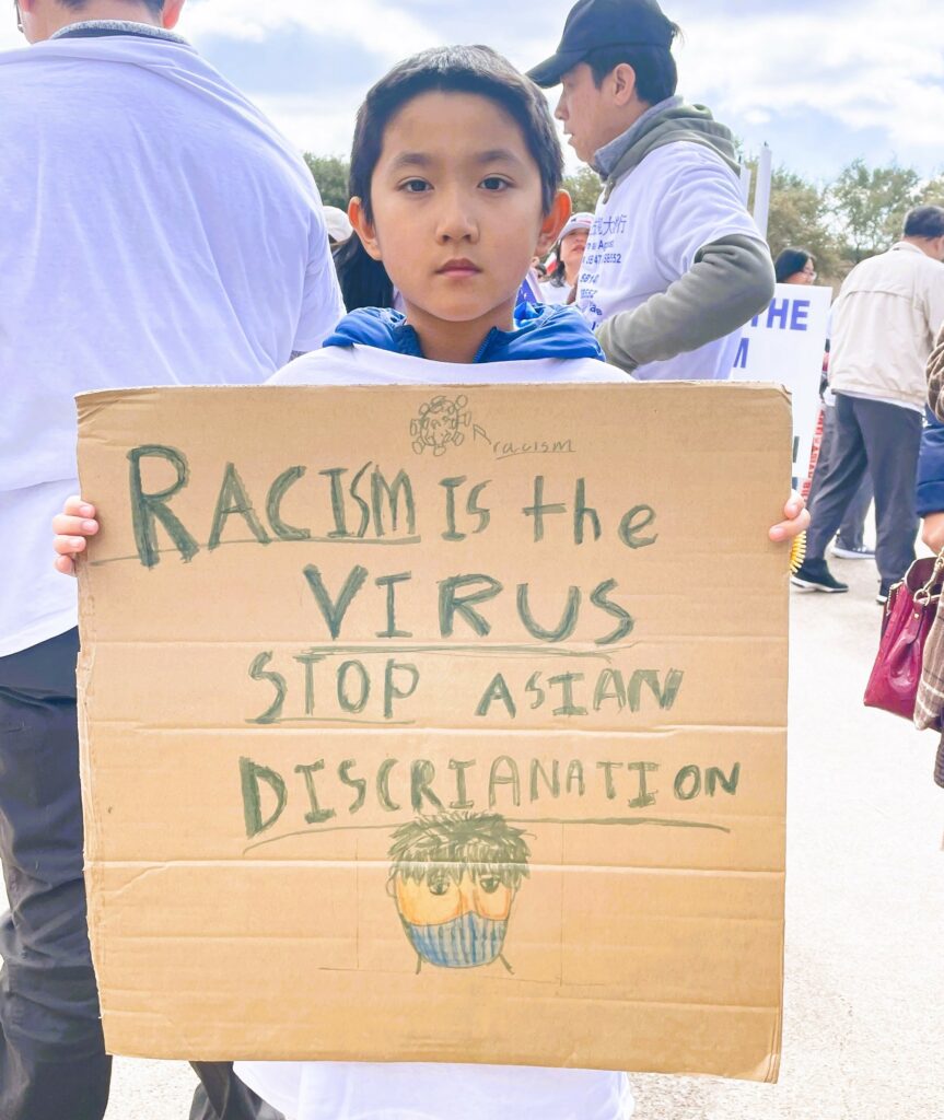 A photo image of a young Chinese American holding up a sign that says Racism is the Virus Stop Asian Discrimination
